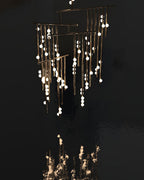 A Clérachic chandelier with brass rods and lit white orbs, set against a near-black backdrop with a bright reflection below.