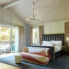 In a bedroom with a pitched timber ceiling, a Clérachic pendant with crossed brass bars hangs above a large bed and a striped bench.