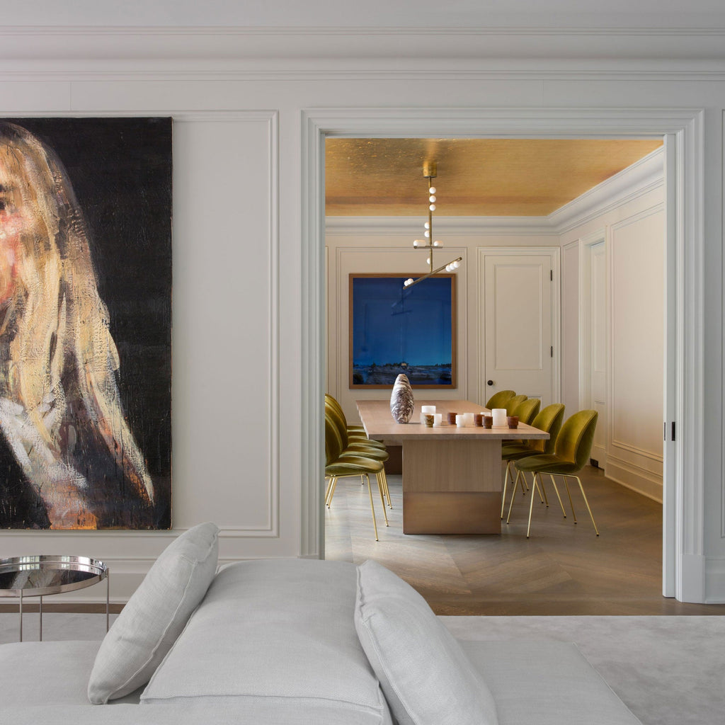 Seen through a wide doorway, a Clérachic pendant hangs in a dining room with olive velvet chairs, past a large portrait in the foreground.