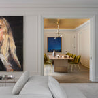 Seen through a wide doorway, a Clérachic pendant hangs in a dining room with olive velvet chairs, past a large portrait in the foreground.