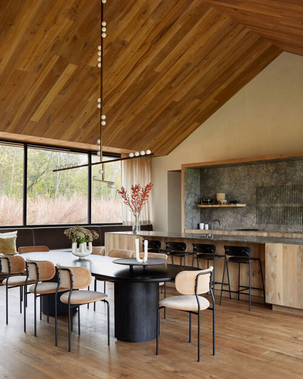 A Clérachic pendant with a long rod and white orbs hangs in a kitchen with a timber ceiling, above a black round dining table.