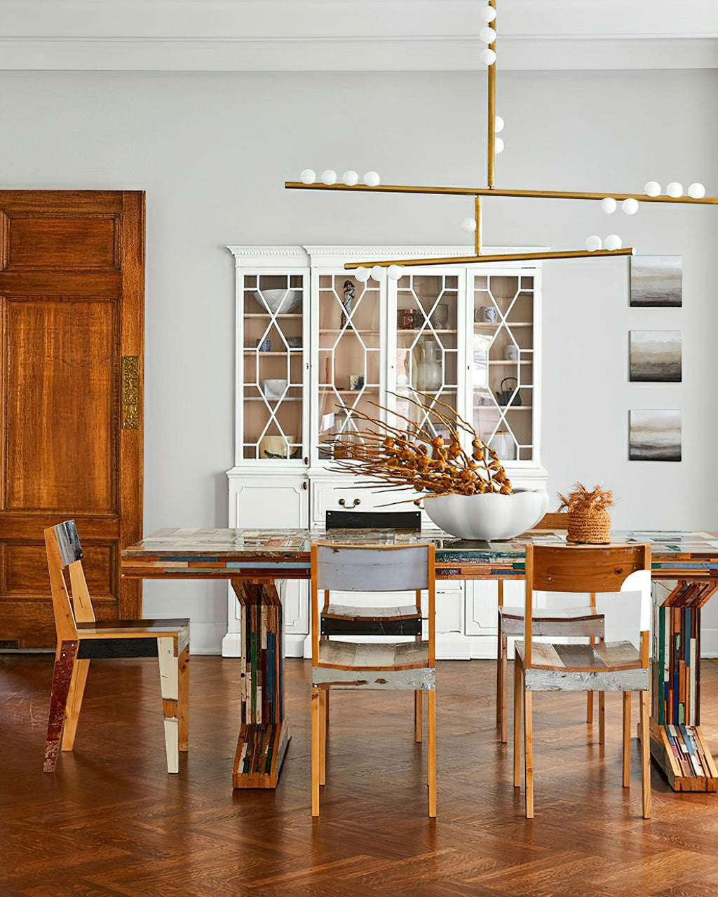 Crossed brass bars of this Clérachic pendant hang over a table with mixed wooden chairs, in front of a white glazed cabinet.