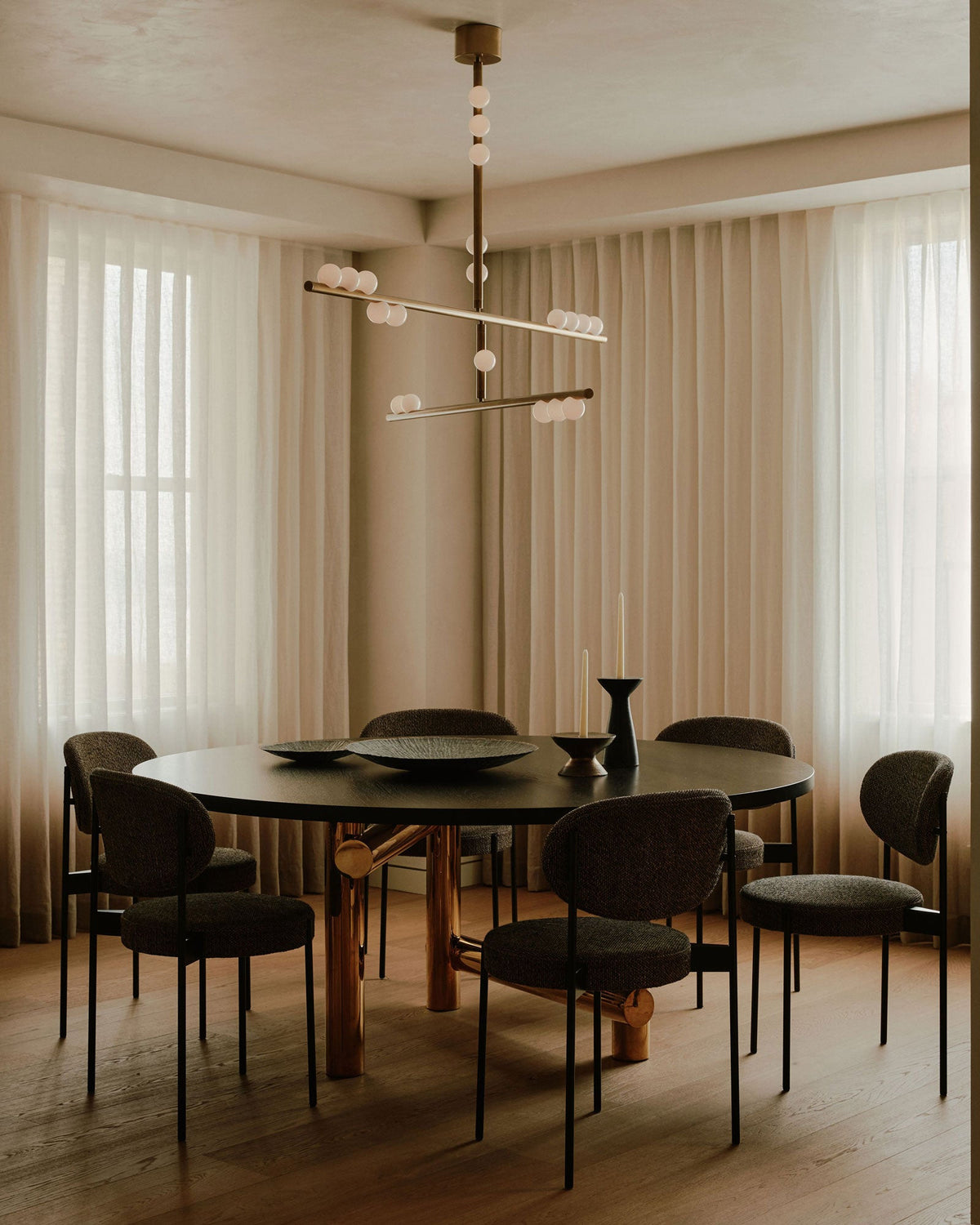 A Clérachic pendant hangs by sheer curtains over a round black dining table, with dark upholstered chairs around it.