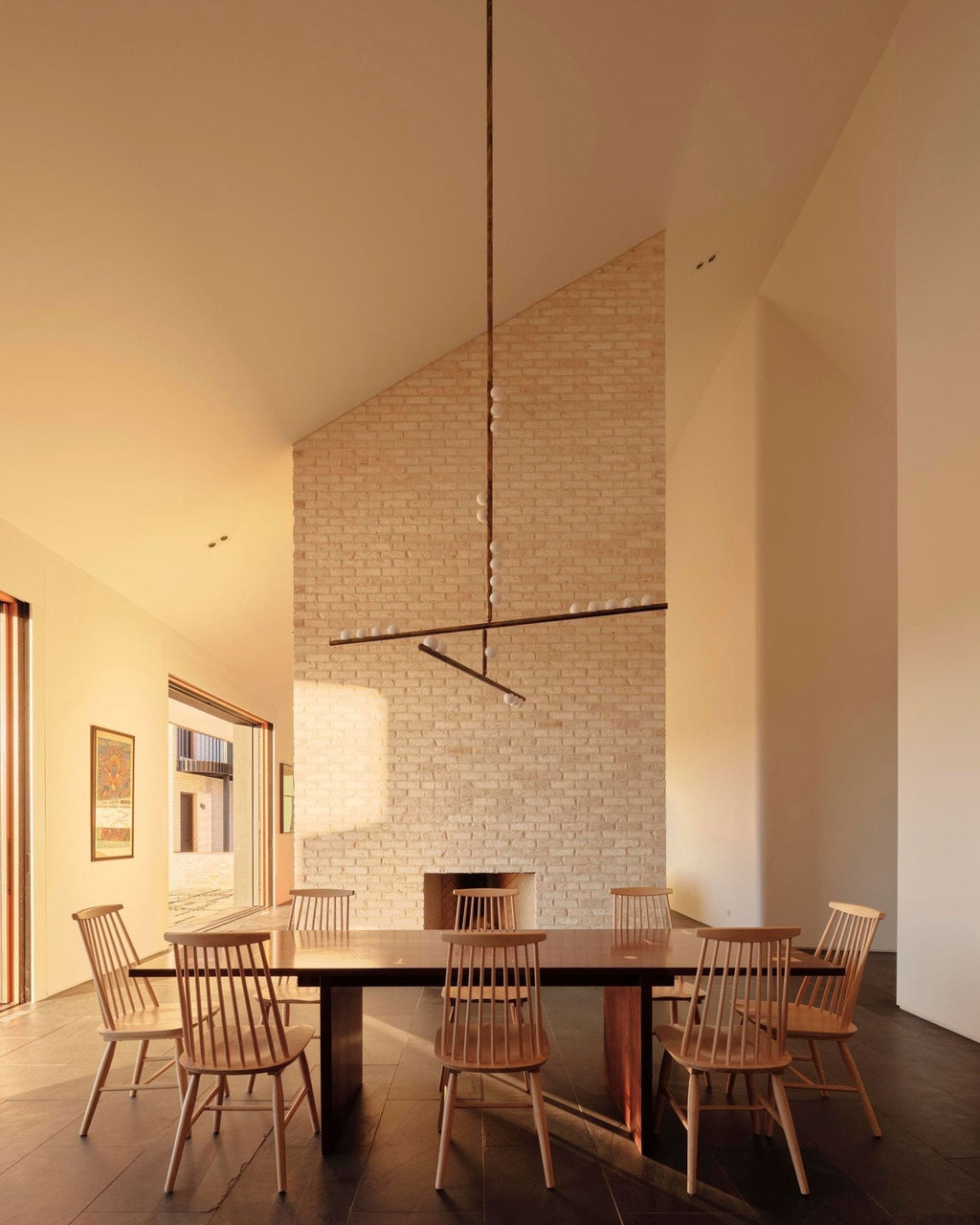 Above a long wooden table with spindle chairs, a Clérachic pendant hangs from a high ceiling against a pale brick wall.