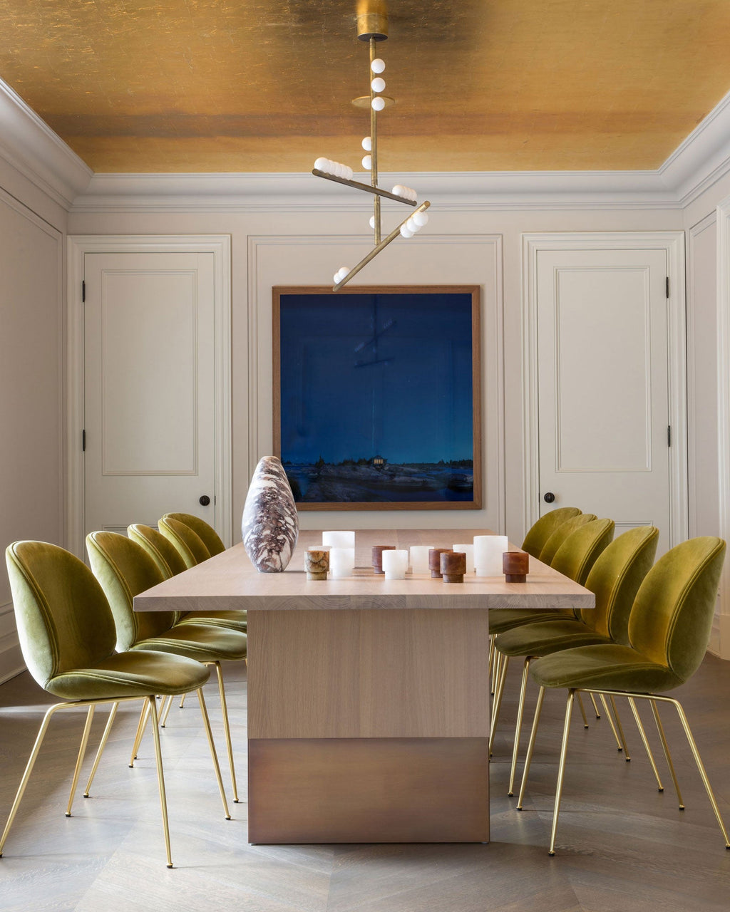 In a dining room with a gold-leaf ceiling, a Clérachic pendant with angled bars hangs above a pale wood table and olive chairs.