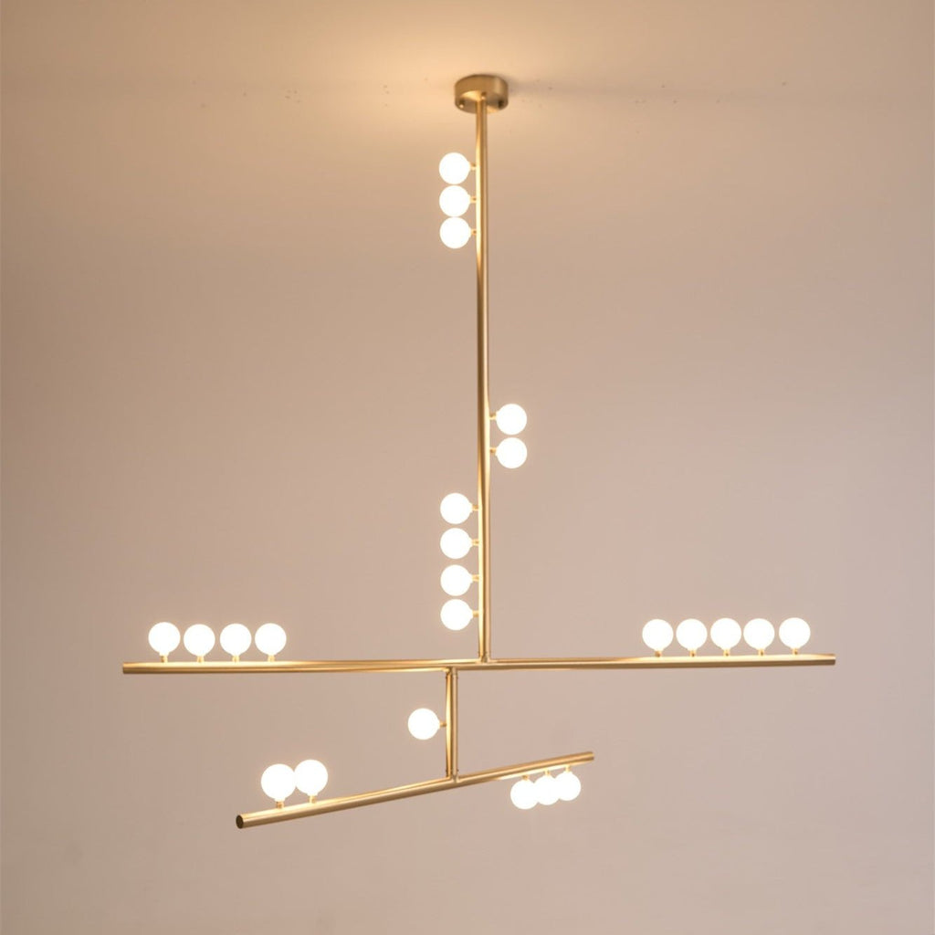 The same Clérachic pendant lit warm, with its brass rod, round canopy and white orbs glowing on a peach-toned backdrop.