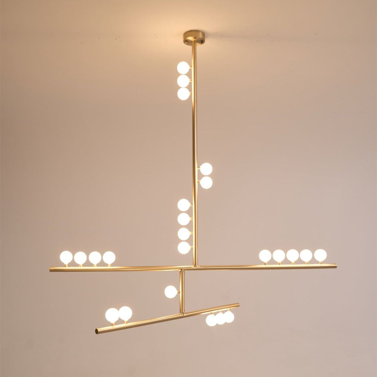 The same Clérachic pendant lit warm, with its brass rod, round canopy and white orbs glowing on a peach-toned backdrop.