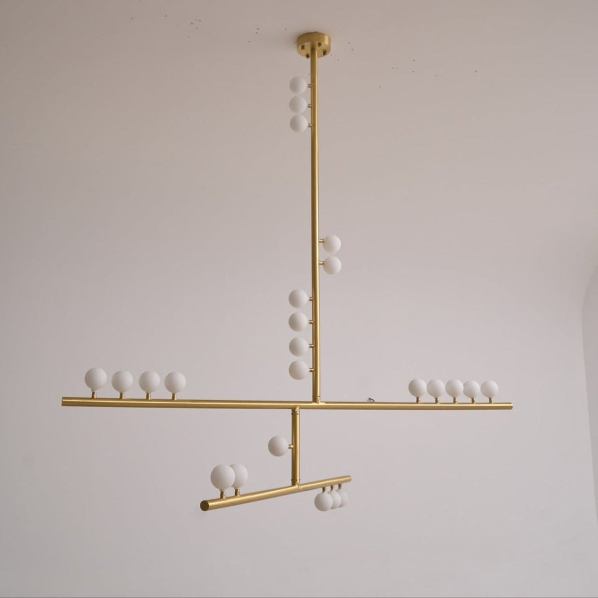 This Clérachic pendant unlit on a pale grey backdrop, with a brass rod, crossed bars and white orbs that look matt.