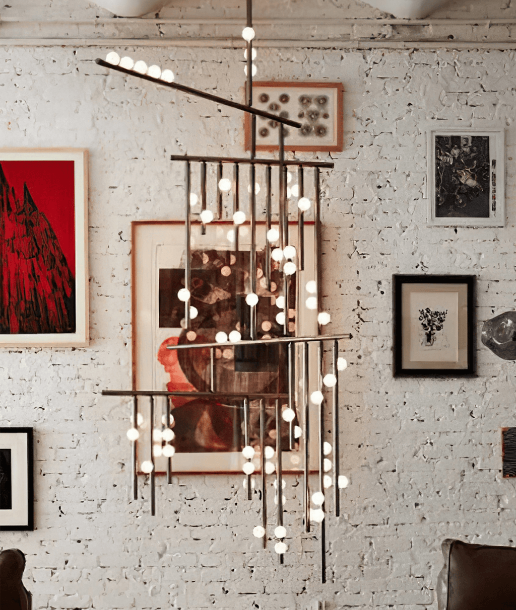 A Clérachic chandelier hangs in a tall brick-walled loft, its dark rods and glowing orbs dropping in front of framed art.