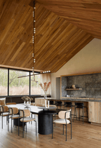 A Clérachic pendant with a long vertical rod hangs under a timber ceiling above a dining table, beside a stone-clad kitchen.