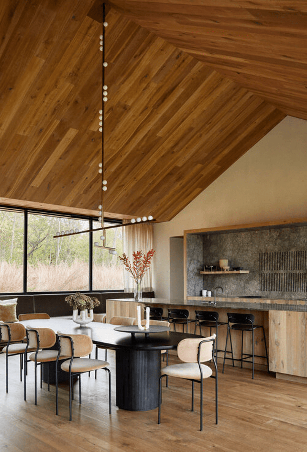 A Clérachic pendant with a long vertical rod hangs under a timber ceiling above a dining table, beside a stone-clad kitchen.