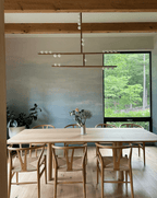 Crossed bars of a Clérachic pendant hang in front of a grey-blue wall above a pale wooden table with wishbone chairs.