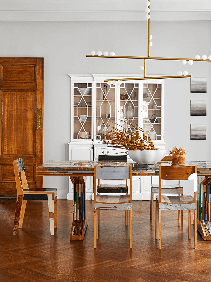 A Clérachic pendant with brass bars hangs over a mixed-wood dining table, in front of a white cabinet with glazed doors.