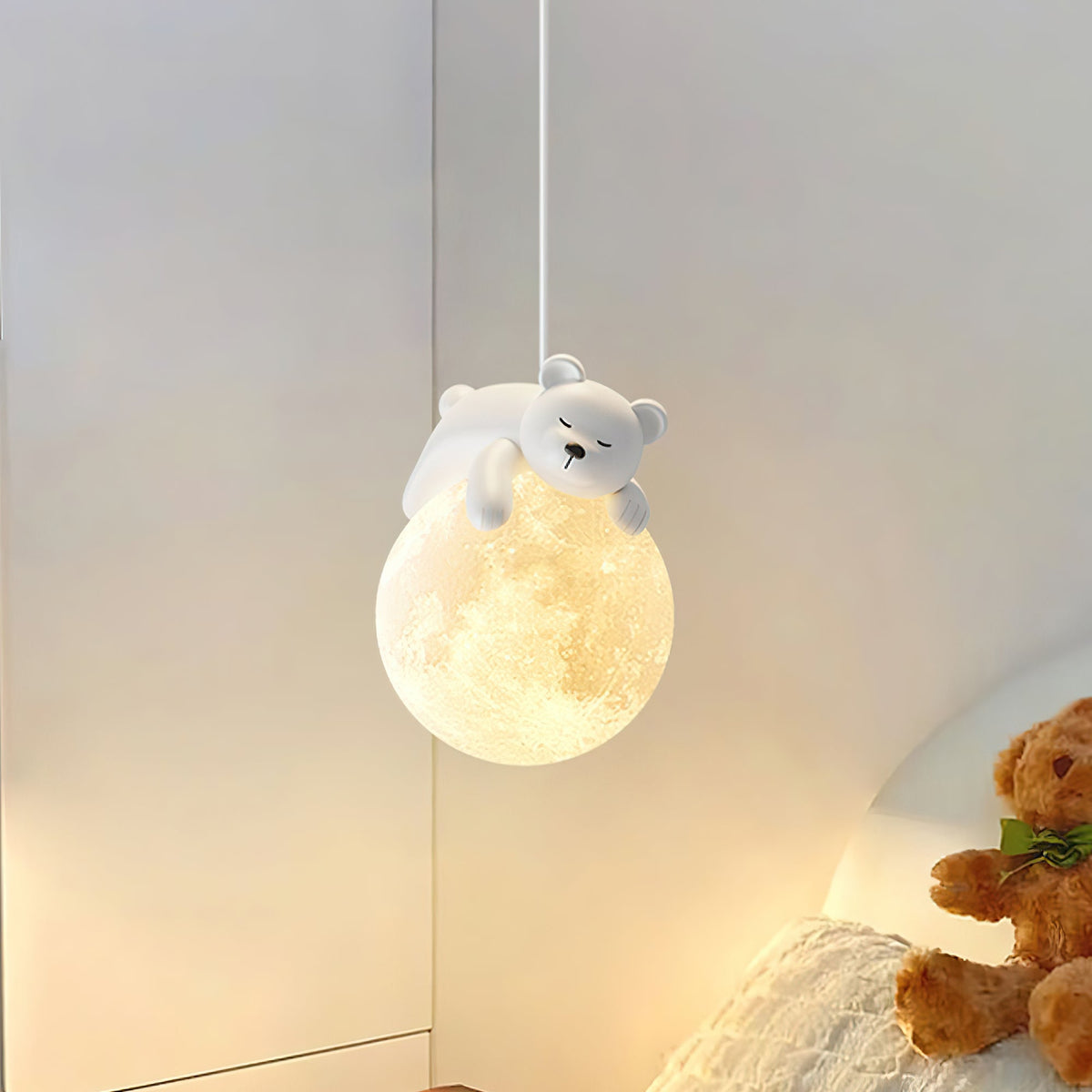 Lit Clérachic pendant with a sleeping bear on a moon-like globe, hanging by a bed with a teddy bear and soft bedding.