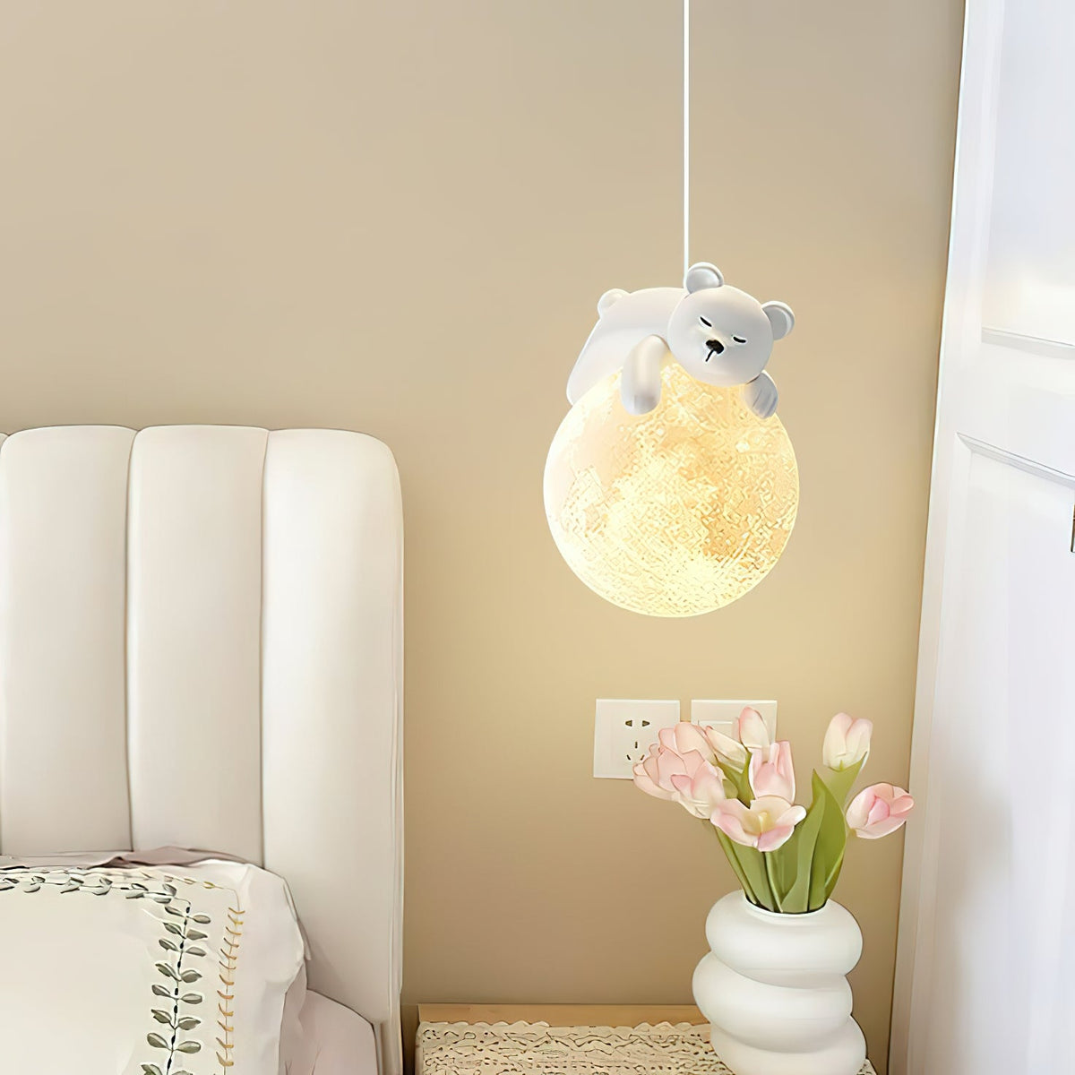 A Clérachic pendant with a sleeping bear on a glowing globe hangs beside a cream padded headboard and a vase of pink tulips.