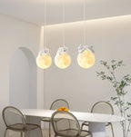 Three Clérachic pendant lights, each with a small sleeping animal on a glowing globe, hang over a white dining table and clear chairs.