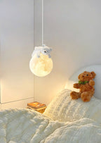 A Clérachic pendant with a bear on a moon globe glows above a bed with textured cream bedding and a teddy bear.
