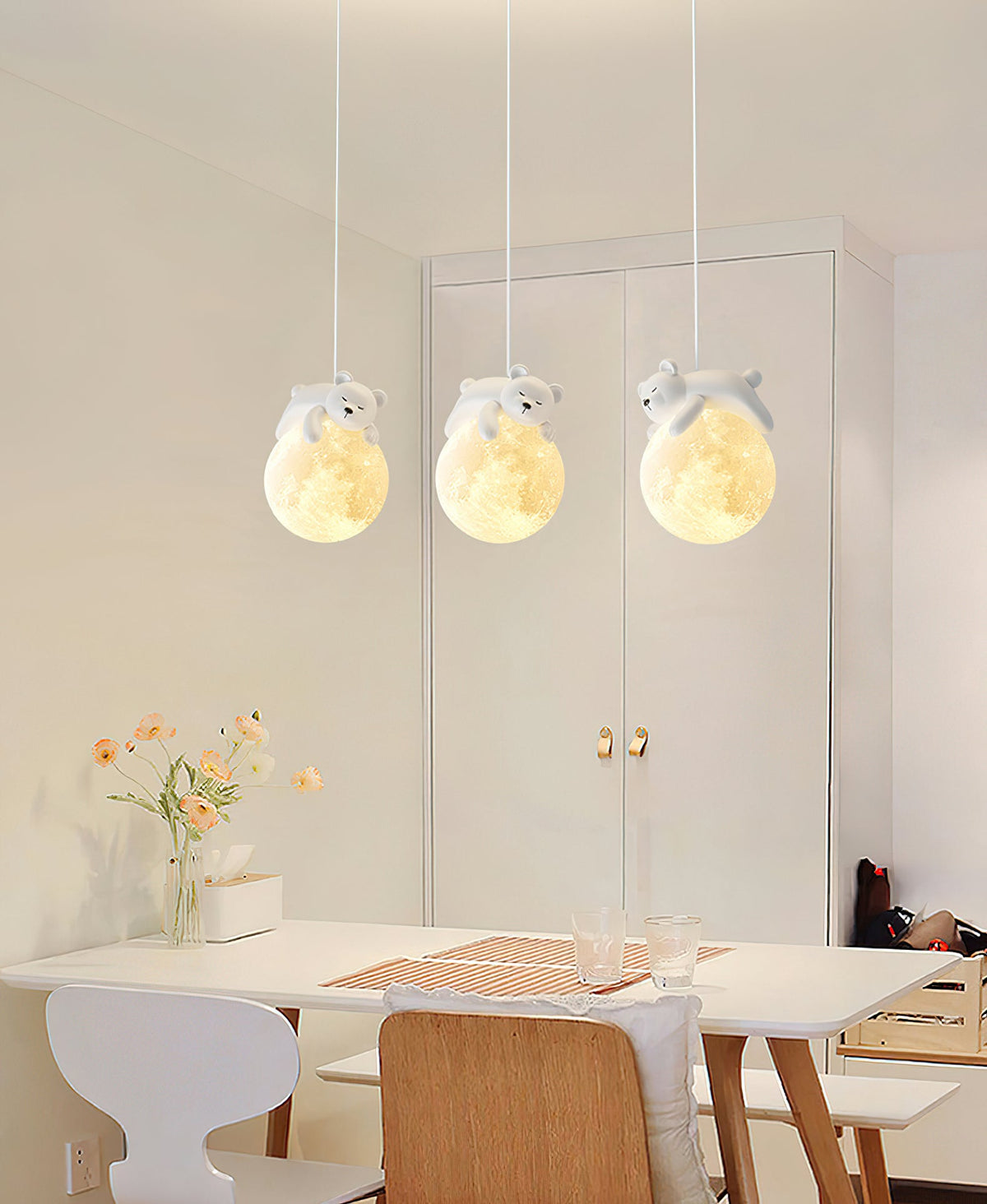 Three Clérachic pendants with sleeping animals on glowing globes hang in front of a white wardrobe, above a table with spring flowers.