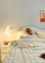 A Clérachic pendant with a sleeping bear glows over a bed with a cloud-shaped headboard, a teddy bear and a cream quilted cover.