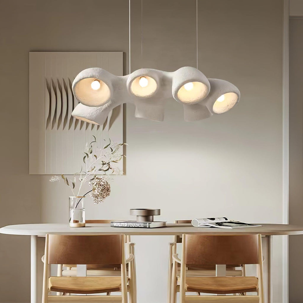 Four-light Clérachic pendant glowing above a pale oval dining table with tan leather chairs, beside a wall panel with a ridged pattern.