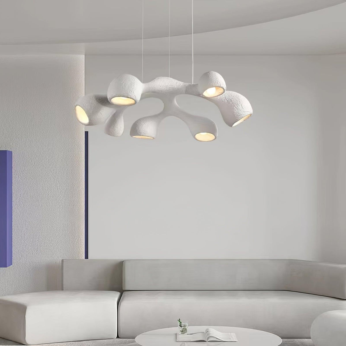 Distant view of a Clérachic pendant with branching white arms over a pale sofa and a round low table, in a grey, curved room.