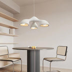 A three-arm Clérachic pendant above a round dark table with cane-backed chairs, next to open wooden shelves on a cream wall.
