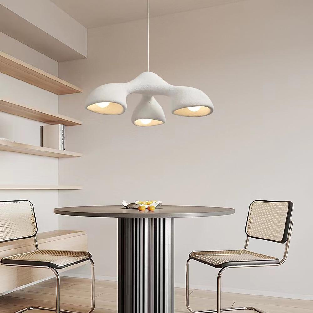 A three-arm Clérachic pendant above a round dark table with cane-backed chairs, next to open wooden shelves on a cream wall.
