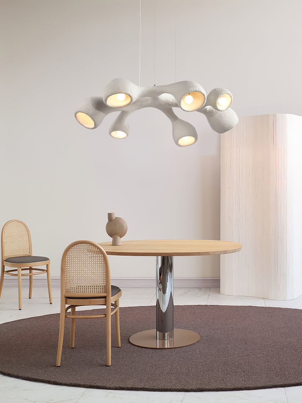 Clérachic pendant above a round wooden table with a steel pedestal, cane chairs and a dark rug, in a pale, airy room.