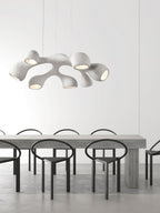 Clérachic pendant hung low above a long grey table with black ring-backed chairs, in a bright minimalist room.