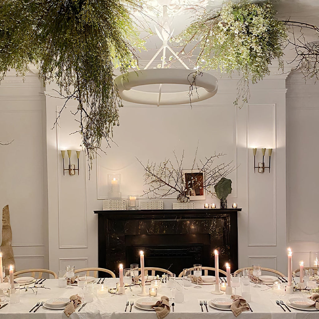 Clérachic ceiling light hanging among trailing greenery over a long dining table set with candles, with a black marble fireplace behind.