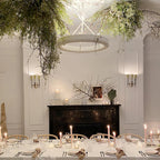 Clérachic ceiling light hanging among trailing greenery over a long dining table set with candles, with a black marble fireplace behind.