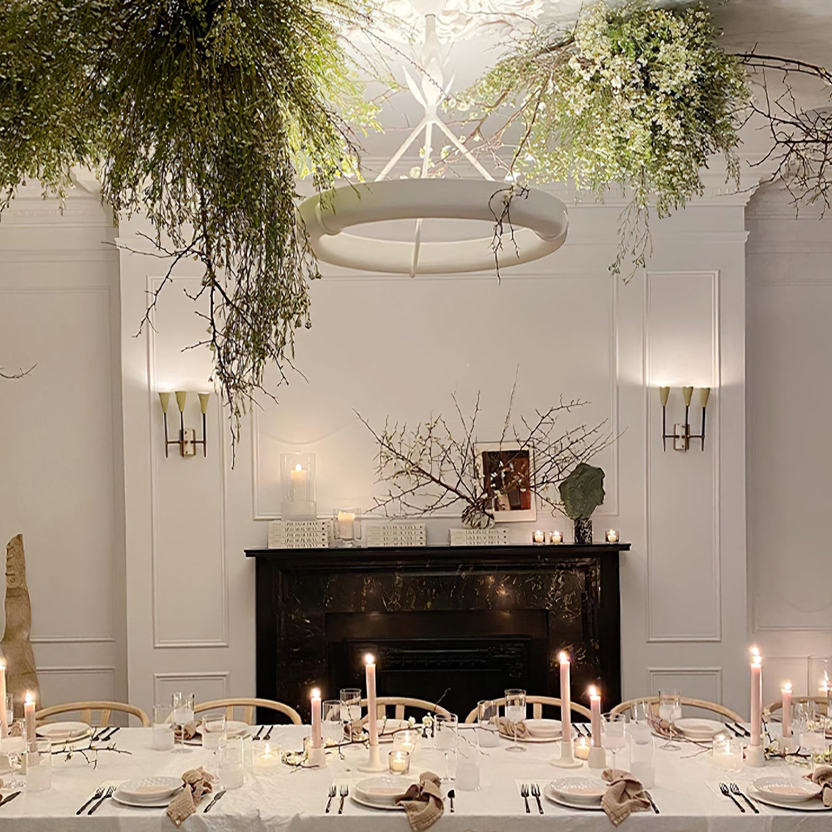 Clérachic ceiling light hanging among trailing greenery over a long dining table set with candles, with a black marble fireplace behind.