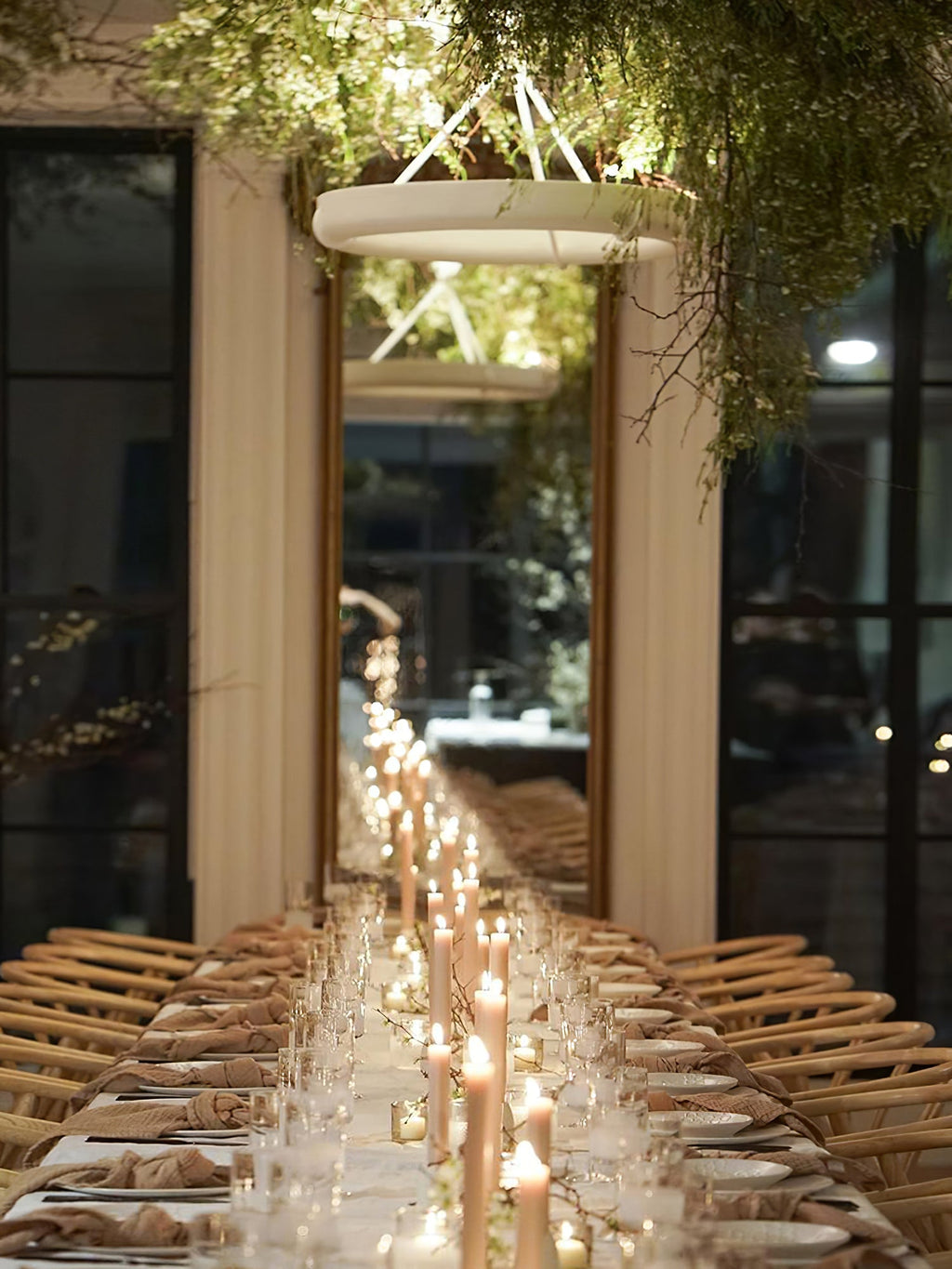 Narrow view down a long candlelit table, with this Clérachic ceiling light hanging among green foliage in a tall window room.