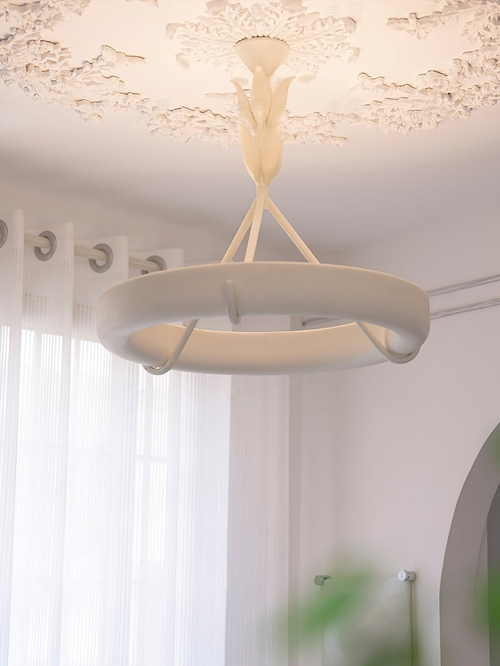 Lit Clérachic ceiling light with a soft glow around the ring, under a plaster rose beside sheer curtains and an arched opening.