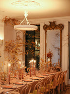 Clérachic ceiling light glowing over a long dining table in warm amber light, with a gilt mirror and autumn branches.