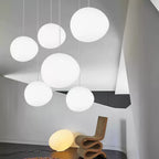Six white Clérachic glass pebble pendants glow in a pale room with a sculptural chair and a stone-shaped floor lamp.