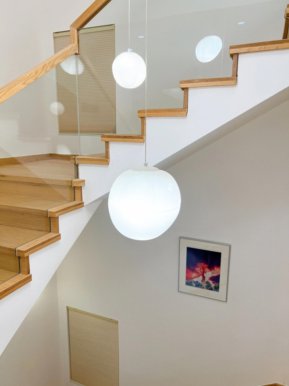A Clérachic white glass pebble pendant glows above a wooden staircase with a glass balustrade and a framed print.
