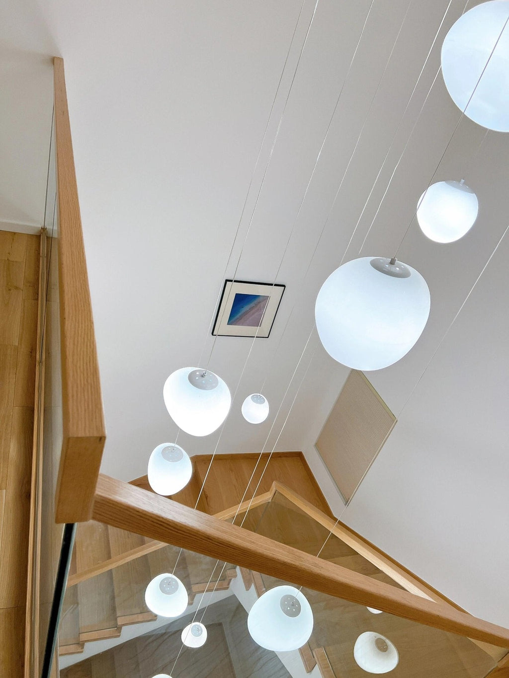 Downward view of a stairwell with oak handrail, hung with Clérachic white glass pebble pendants of several sizes.