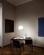 Single white Clérachic glass pebble pendant on a thin cord above a desk and chair in a calm, dark-walled study.