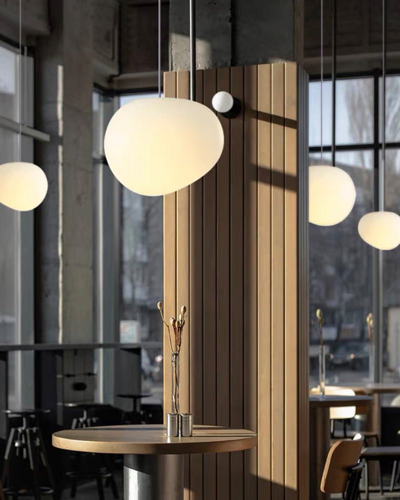 A Clérachic white glass pebble pendant glows warm by a timber partition in a café with tall industrial windows.