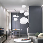 A group of white Clérachic glass pebble pendants hangs above a round white table and sofa in a grey-panelled room.