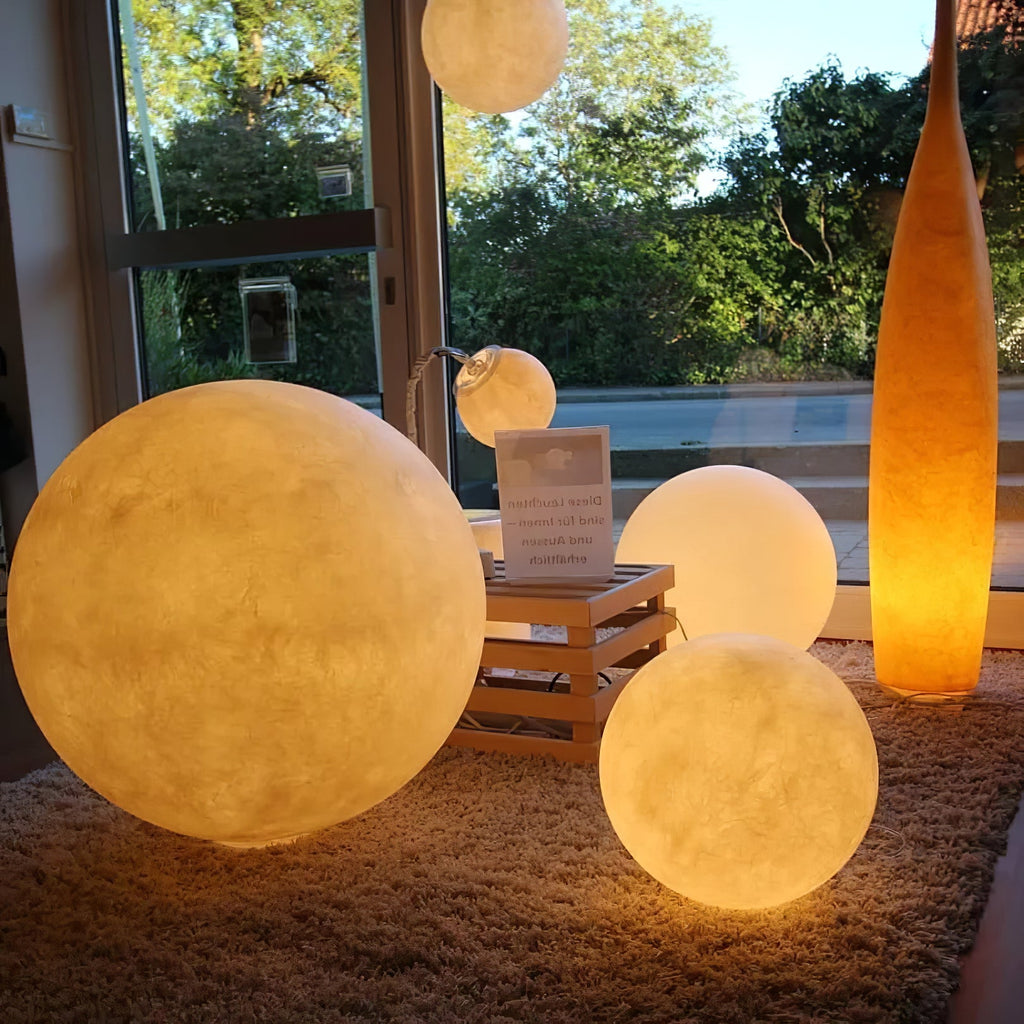 Four glowing Clérachic spheres in different sizes on a shaggy rug, lit warm amber beside a window onto a green garden.