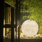 A large white Clérachic sphere lamp glowing among white pebbles and bamboo leaves, seen through dark window frames.