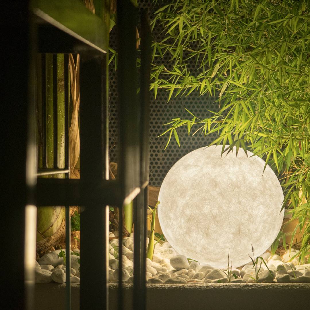 A large white Clérachic sphere lamp glowing among white pebbles and bamboo leaves, seen through dark window frames.