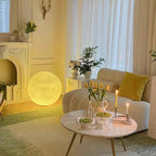 Small warm-glowing Clérachic sphere lamp on the floor by a white fireplace, beside a cream sofa and a round marble coffee table.