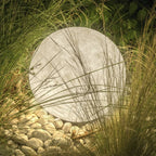 Textured Clérachic sphere lamp lit at dusk, half hidden by tall grasses and resting on a bed of white pebbles.