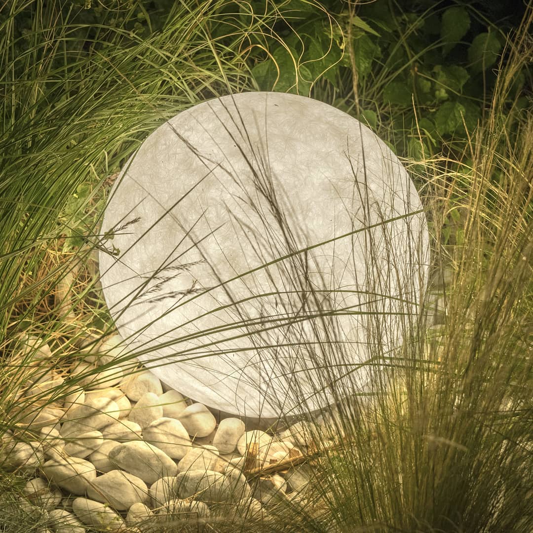 Textured Clérachic sphere lamp lit at dusk, half hidden by tall grasses and resting on a bed of white pebbles.