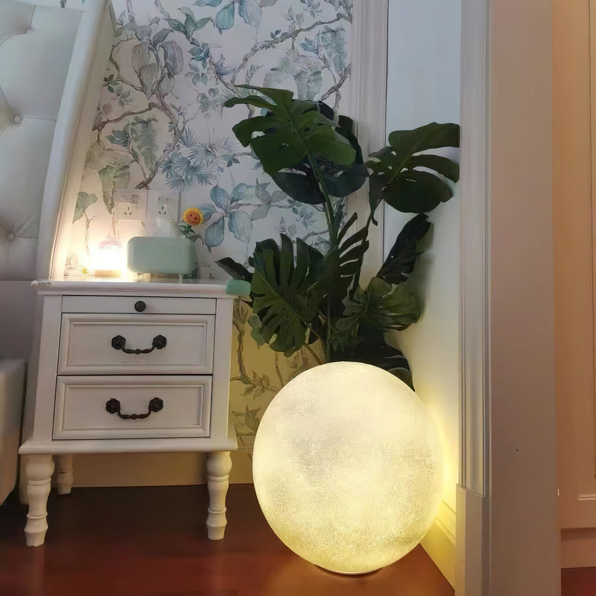 Glowing Clérachic sphere lamp on a wooden floor in a bedroom corner, next to a white two-drawer nightstand and a large leafy plant.
