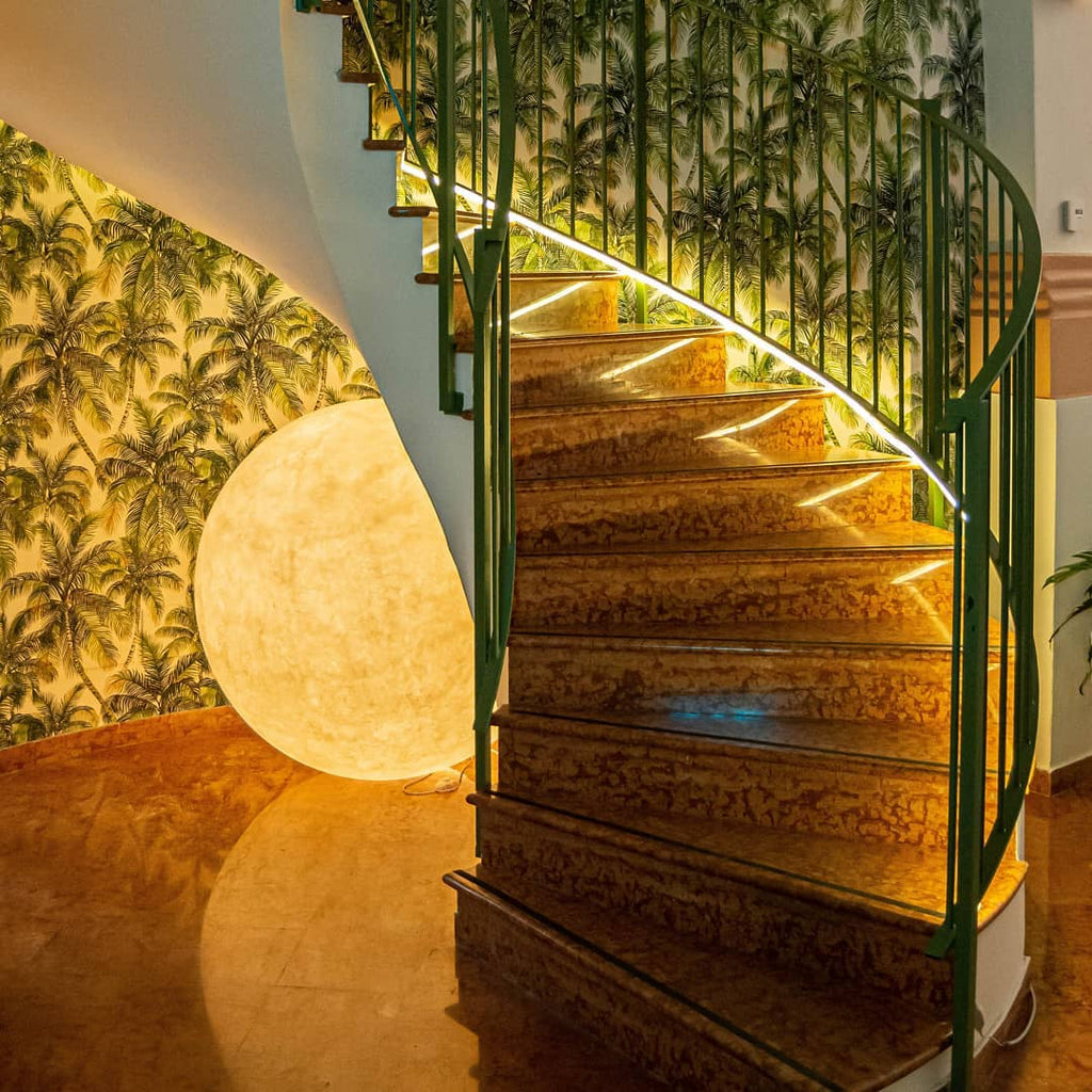 A softly lit Clérachic sphere lamp tucked under a curved staircase with a green railing and palm-print wallpaper.