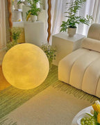 Yellow-lit Clérachic sphere lamp on a green woven rug beside a cream curved sofa, with a pale plinth of plants behind it.